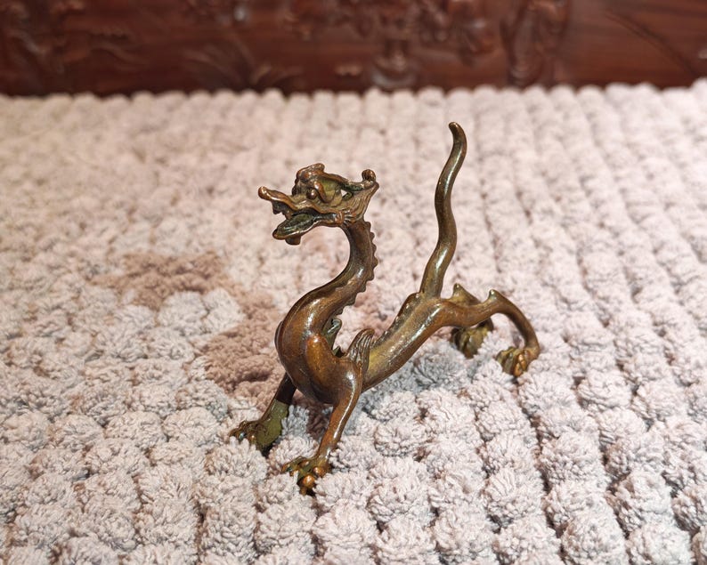 K&ouml;nnte beinhalten: Eine bronzefarbene Drachenfigur mit detailliertem Design. Der Drache ist mit erhobenem Kopf und gebogenem K&ouml;rper dargestellt. Die Figur besteht aus einem metallischen Material und hat ein verwittertes Aussehen. Der Drache ist ein dekoratives Objekt.