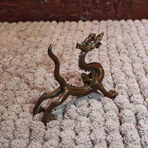 K&ouml;nnte beinhalten: Eine bronzefarbene Drachenfigur mit gew&ouml;lbtem K&ouml;rper, detailliertem Kopf und ausgestreckten Beinen. Der Drache hat einen langen, gebogenen Schwanz und ist auf einer strukturierten Oberfl&auml;che platziert. Dieser Dekorationsartikel ist wahrscheinlich ein Sammlerst&uuml;ck oder ein Einrichtungsgegenstand.