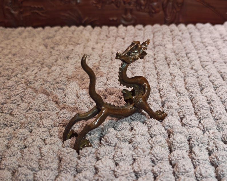K&ouml;nnte beinhalten: Eine bronzefarbene Drachenfigur mit gew&ouml;lbtem K&ouml;rper, detailliertem Kopf und ausgestreckten Beinen. Der Drache hat einen langen, gebogenen Schwanz und ist auf einer strukturierten Oberfl&auml;che platziert. Dieser Dekorationsartikel ist wahrscheinlich ein Sammlerst&uuml;ck oder ein Einrichtungsgegenstand.