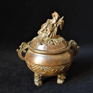 May include: A bronze-colored incense burner with a dragon figure on the lid. The burner has a round body with ornate carvings, three feet, and handles. The lid has decorative cutouts.