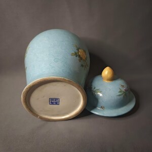 May include: A light blue ceramic vase with a matching lid. The vase features floral designs and a textured pattern. The lid has a yellow finial. The base of the vase has a blue square stamp.
