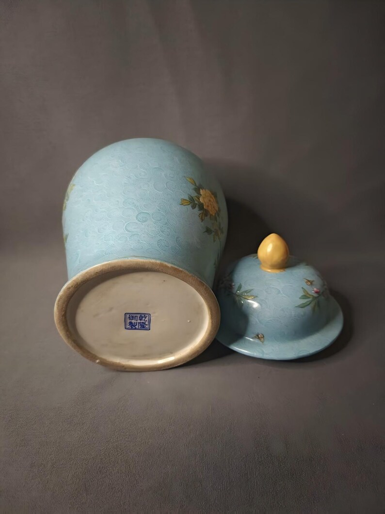 May include: A light blue ceramic vase with a matching lid. The vase features floral designs and a textured pattern. The lid has a yellow finial. The base of the vase has a blue square stamp.