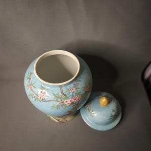 May include: A light blue ceramic jar with a matching lid. The jar and lid are decorated with a floral design featuring pink blossoms and green leaves. The lid has a yellow knob.