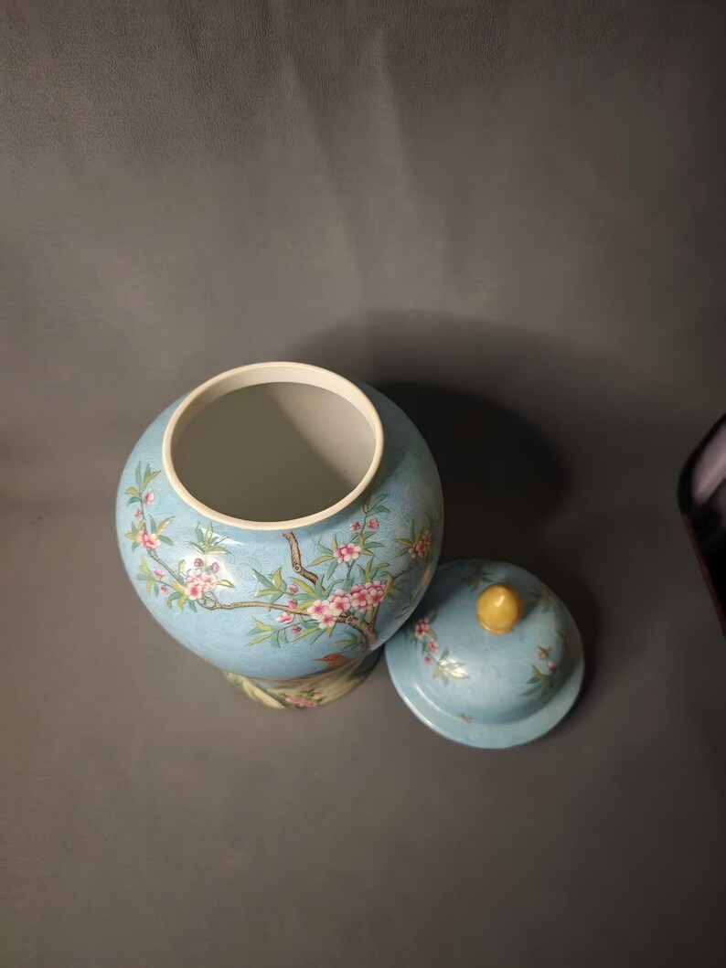 May include: A light blue ceramic jar with a matching lid. The jar and lid are decorated with a floral design featuring pink blossoms and green leaves. The lid has a yellow knob.