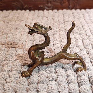 K&ouml;nnte beinhalten: Bronze-farbene Drachenfigur mit aufwendigen Details. Der Drache hat einen langen, geschwungenen K&ouml;rper, vier Beine und einen detaillierten Kopf mit Krone. Die Figur ist aus Metall und ein Dekorationsartikel.