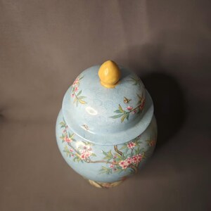 May include: A light blue ceramic jar with a lid, adorned with a floral design featuring pink blossoms and green leaves. The lid is topped with a yellow finial. The jar has a bulbous shape and is likely used for decorative purposes.