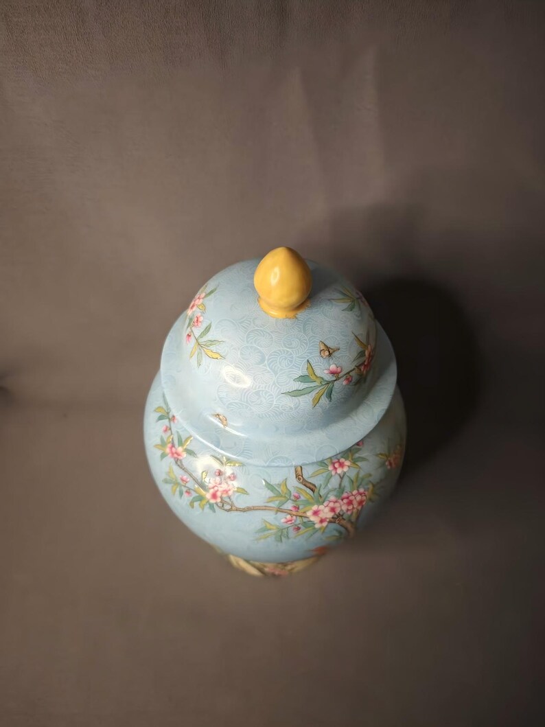May include: A light blue ceramic jar with a lid, adorned with a floral design featuring pink blossoms and green leaves. The lid is topped with a yellow finial. The jar has a bulbous shape and is likely used for decorative purposes.