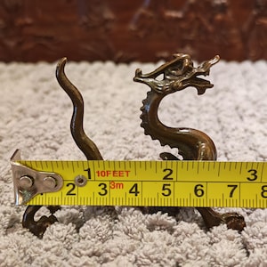 K&ouml;nnte beinhalten: Bronze-Drachenfigur, ca. 7,6 cm lang, mit aufwendigen Details. Der Drache ist mit gebogenem K&ouml;rper und erhobenem Kopf dargestellt. Ein gelbes Ma&szlig;band liegt neben dem Drachen.