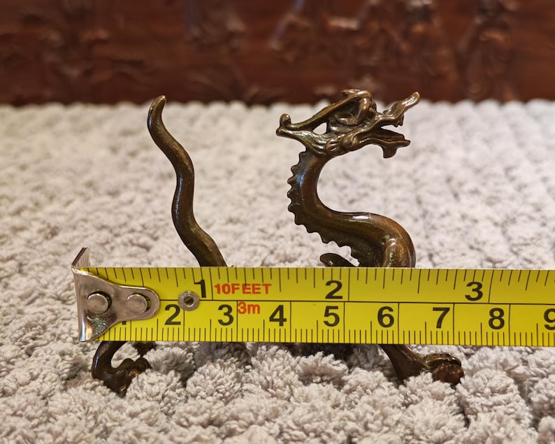 K&ouml;nnte beinhalten: Bronze-Drachenfigur, ca. 7,6 cm lang, mit aufwendigen Details. Der Drache ist mit gebogenem K&ouml;rper und erhobenem Kopf dargestellt. Ein gelbes Ma&szlig;band liegt neben dem Drachen.
