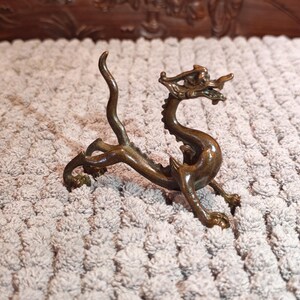 K&ouml;nnte beinhalten: Eine bronzene Drachenfigur mit aufwendigen Details. Der Drache ist in einer dynamischen Pose dargestellt, mit gew&ouml;lbtem K&ouml;rper und erhobenem Kopf, der ein detailliertes Gesicht und Schuppen zeigt. Auf hellem, strukturiertem Hintergrund.