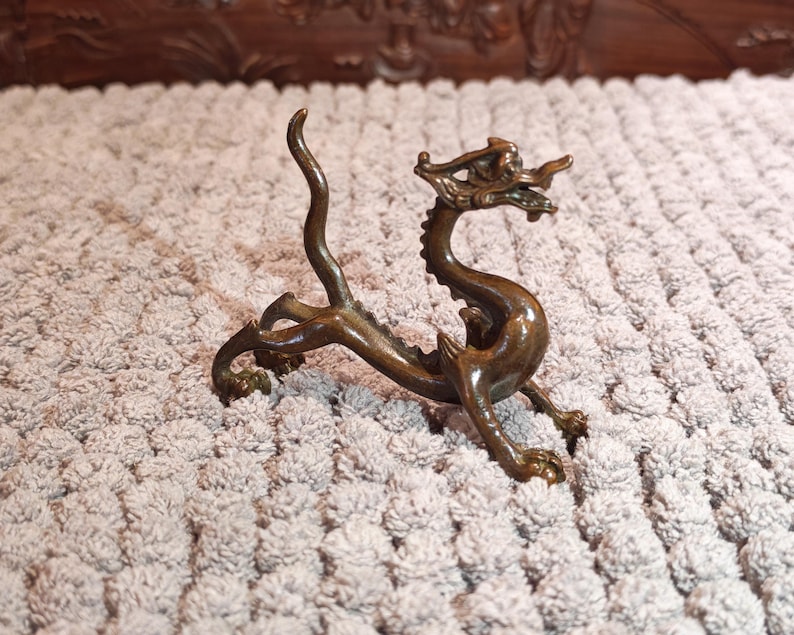 K&ouml;nnte beinhalten: Eine bronzene Drachenfigur mit aufwendigen Details. Der Drache ist in einer dynamischen Pose dargestellt, mit gew&ouml;lbtem K&ouml;rper und erhobenem Kopf, der ein detailliertes Gesicht und Schuppen zeigt. Auf hellem, strukturiertem Hintergrund.