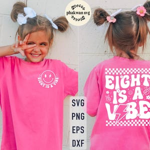 May include: A bright pink t-shirt with the text "EIGHT IS A VIBE" in white. The front of the shirt features a smiley face graphic. The back of the shirt has a checkered pattern and additional graphics.