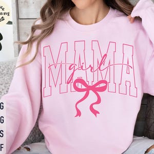 Könnte beinhalten: Hellrosa Sweatshirt mit dem Wort "MAMA" in großen Blockbuchstaben. Das Wort "girl" ist in Schreibschrift über der Mitte des Wortes "MAMA" geschrieben. Darunter befindet sich eine rosa Schleife.