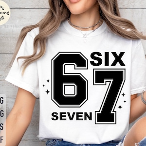 Puede incluir: Camiseta blanca con un diseño gráfico negro que presenta el número "67" y las palabras "SIX" y "SEVEN". El diseño tiene un estilo universitario con una fuente en negrita y acentos de estrellas. Es una prenda informal.