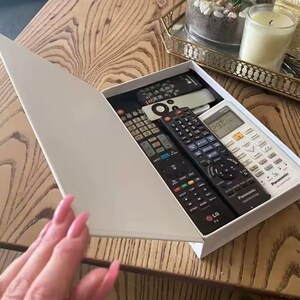 May include: A white box with a lid, containing several black remote controls. The remotes are of various sizes and brands, including LG and Panasonic. The box is on a wooden table.