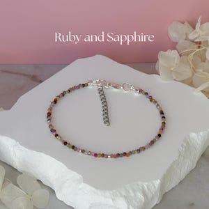 May include: A delicate bracelet with small, colourful beads, possibly gemstones, strung together with a silver-toned clasp and extender chain. The text "Ruby and Sapphire" is displayed above the bracelet against a pink background.