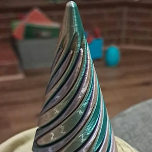 May include: A 3D-printed, cone-shaped object with a spiral design. The object features a gradient of colours, including teal, silver, and purple. The design has a textured, ridged surface.