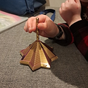 以下が含まれることがあります： らせん状のハンドルが付いた、金と紫色の星型のオーナメント。オーナメントは、星型のベースとピラミッドのような構造を持つ、多層構造のデザインです。オーナメントは人が持っています。