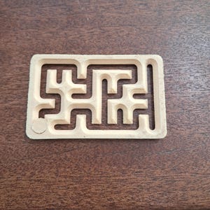 May include: A light beige, rectangular maze puzzle with a complex path design. The puzzle is made of a solid material and sits on a dark brown wooden surface. The maze features a circular starting point.