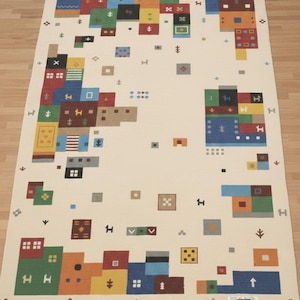 May include: Rectangular off-white rug with a colorful geometric design. The rug features blocks of red, blue, green, yellow, and orange, with various symbols and shapes. The rug is placed on a wooden floor.