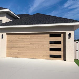May include: A light-colored wooden garage door with four rectangular black windows. The door is part of a modern house with a dark roof and white trim. The driveway is concrete, and the sky is blue.