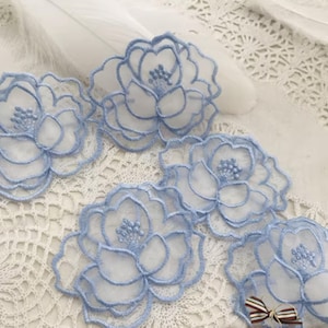 May include: Close-up of several light blue embroidered flower appliques. The flowers have a delicate, open design with detailed stitching and a soft, airy appearance. The appliques are arranged on a white lace surface, suggesting a craft or sewing project.
