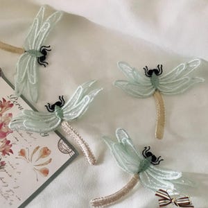 May include: Four delicate dragonfly embellishments. Each dragonfly features pale blue wings, a black body, and a beige stem. One dragonfly rests near a postcard with floral and butterfly illustrations. The dragonflies are arranged on a white surface.