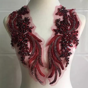 May include: A burgundy and silver beaded applique with a floral and feather design. The applique features a sheer, light pink mesh backing. The design includes clusters of dark red rhinestones and silver beads.