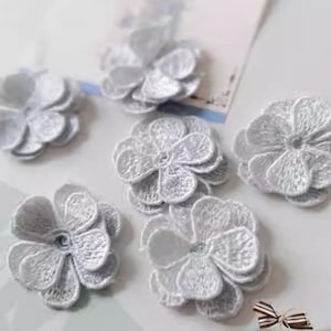 Può includere: Una collezione di applicazioni floreali bianche ricamate. Ogni fiore ha più petali e un aspetto dettagliato e strutturato. Le applicazioni sono disposte su una superficie chiara, con un piccolo nastro visibile nell'angolo.