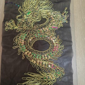 May include: A gold-colored dragon embroidered on a dark gray mesh fabric. The dragon design features intricate details with green, pink, and gold sequins. The dragon's body is outlined with gold thread, creating a textured and ornate appearance.