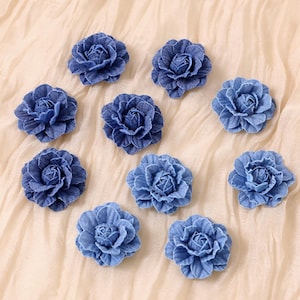 May include: A collection of ten denim blue fabric flower embellishments. Each flower has multiple layers of petals, creating a realistic rose-like appearance. The flowers are arranged on a light beige fabric background.