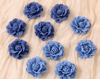 Pode incluir: Uma coleção de dez enfeites de flores de tecido azul denim. Cada flor tem múltiplas camadas de pétalas, criando uma aparência realista de rosa. As flores estão dispostas sobre um fundo de tecido bege claro.