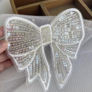May include: A white, sequined bow applique. The bow is intricately detailed with small, shimmering sequins and beads, creating a delicate, textured appearance. The bow's outline is defined by a thin, white border.