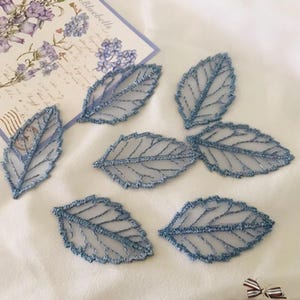 May include: Delicate blue leaf-shaped appliques with intricate vein detailing. The appliques are made of a sheer, shimmering fabric, and are scattered on a white surface. A floral card is in the background.