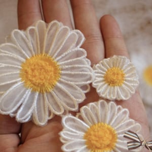 Puede incluir: Primer plano de varios apliques de flores de margarita blancas con centros amarillos. Los apliques están hechos de una tela delicada y transparente y están dispuestos sobre una mano. Se ve un pequeño lazo decorativo.