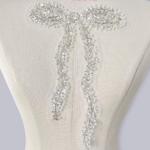 May include: A silver sequin and bead bow applique. The bow is attached to a white fabric form. The bow has two long, flowing tails. The sequins and beads are arranged to create a sparkling effect.