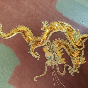 May include: A golden dragon applique with intricate detailing, including sequins and embroidery. The dragon features a long, serpentine body and is set against a maroon and green background. The dragon's eyes are blue.