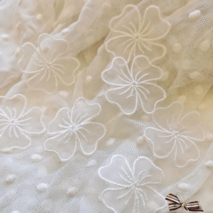 May include: Close-up of a white, delicate fabric with embroidered floral details. The fabric has a dotted texture and features several large, white, embroidered flowers. A small, striped bow is visible in the bottom right corner.
