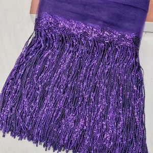 May include: Close-up of a length of purple fringe trim. The trim features a beaded top section and long, dangling strands of fringe. The color is a deep, vibrant purple, with a slight sheen. This decorative trim is suitable for various craft and design projects.