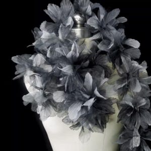 May include: A gray floral accessory, possibly a scarf or collar, displayed on a mannequin. The flowers are made of layered, sheer fabric in shades of gray and white, creating a delicate, textured appearance. The background is black.