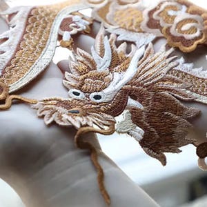 May include: Detailed embroidered dragon patch in brown, gold, and white. The dragon design features intricate detailing, with a long body and detailed head. The patch is suitable for clothing or craft projects. The measurements are in centimeters.