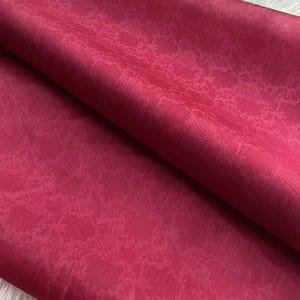 May include: Close-up of a folded piece of fabric in a deep red hue. The fabric has a subtle, repeating floral pattern across its surface. The folds create shadows and highlights, adding depth to the image.