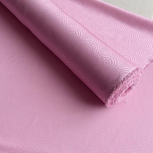 Puede incluir: Un rollo de tela rosa con un patrón repetitivo en forma de olas. La tela es de color rosa liso con un brillo sutil. El patrón es de un tono rosa más claro, creando un efecto texturizado. La tela está enrollada, revelando el borde del material.