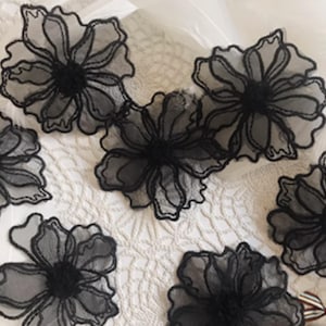 Könnte beinhalten: Mehrere schwarze Spitzenblumen-Applikationen sind auf einer weißen Oberfläche angeordnet. Jede Blume hat detaillierte Blütenblätter und ein dunkles Zentrum, was ein zartes und elegantes Aussehen erzeugt. Die Spitze verleiht Textur.