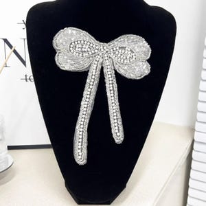 May include: A silver sequined bow brooch with a rhinestone centre and dangling tails. The bow is displayed on a black mannequin. The brooch is a decorative accessory.