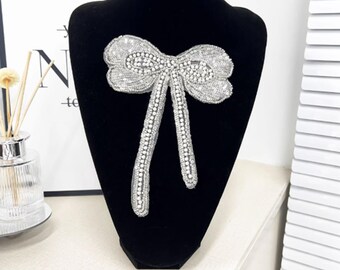 Könnte beinhalten: Silberne Pailletten-Schleifenbrosche mit Strass-Mittelteil und herabhängenden Enden. Die Schleife wird auf einer schwarzen Schaufensterpuppe präsentiert. Ein dekoratives Accessoire.