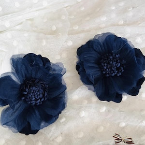 May include: Two navy blue fabric flower hair accessories. Each flower has multiple layers of petals and a textured center. The flowers are set against a white, dotted fabric background. The flowers appear to be handmade.