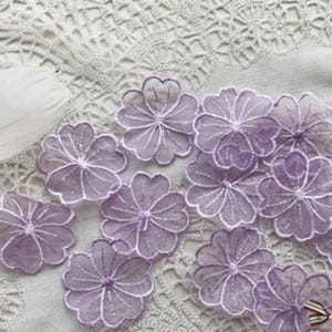 Op de afbeelding: Een verzameling delicate, lavendelkleurige bloemenapplicaties. Elke bloem heeft een gedetailleerde, wit gestikte omtrek en een subtiele, glinsterende textuur. De applicaties zijn verspreid op een getextureerd, gebroken witte kanten oppervlak, met een witte veer op de achtergrond.