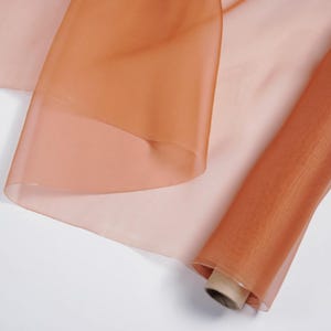 Könnte beinhalten: Eine Rolle transparenten, orangebraunen Stoffes. Der Stoff ist teilweise abgerollt und zeigt eine hellere, pfirsichfarbene Schicht darunter. Das Material wirkt leicht und zart, geeignet für verschiedene Bastelprojekte.