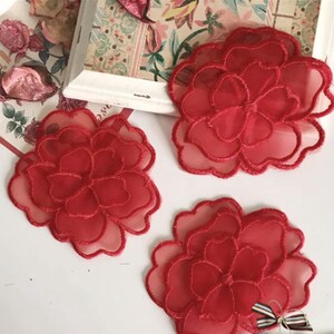 Puede incluir: Tres apliques de flores rojas de tela transparente en capas. Cada flor tiene múltiples pétalos con un contorno delicado cosido. Las flores están dispuestas sobre una superficie blanca con un fondo decorativo y un pequeño lazo.
